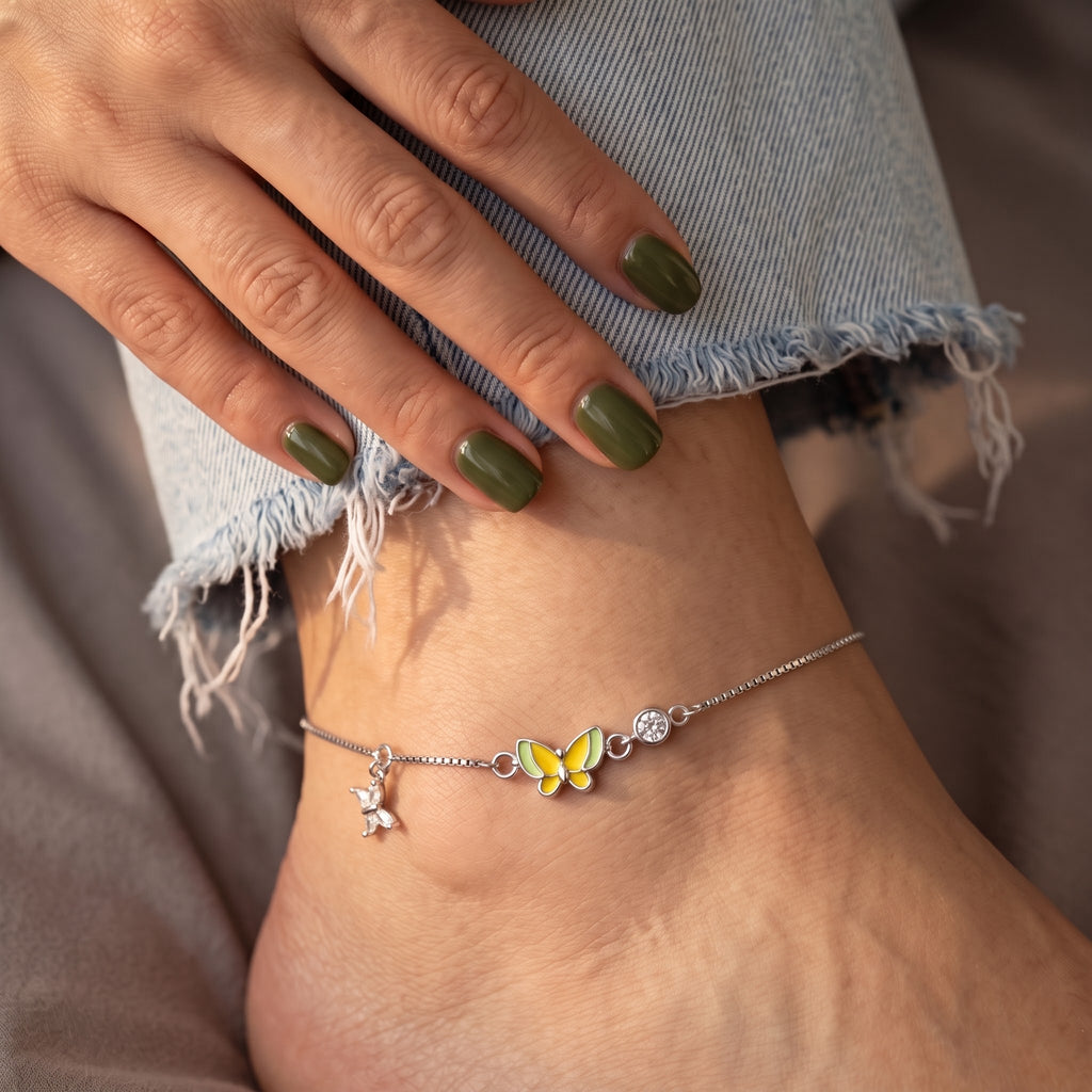 A delicate silver anklet with a yellow and green butterfly charm, a small silver bird charm, and a clear crystal bead, shown worn on a foot with light blue frayed denim jeans.