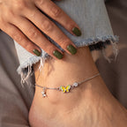 A delicate silver anklet with a yellow and green butterfly charm, a small silver bird charm, and a clear crystal bead, shown worn on a foot with light blue frayed denim jeans.