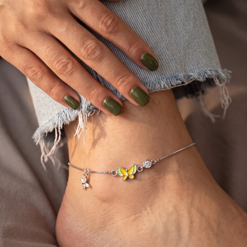 A delicate silver anklet with a yellow and green butterfly charm, a small silver bird charm, and a clear crystal bead, shown worn on a foot with light blue frayed denim jeans.